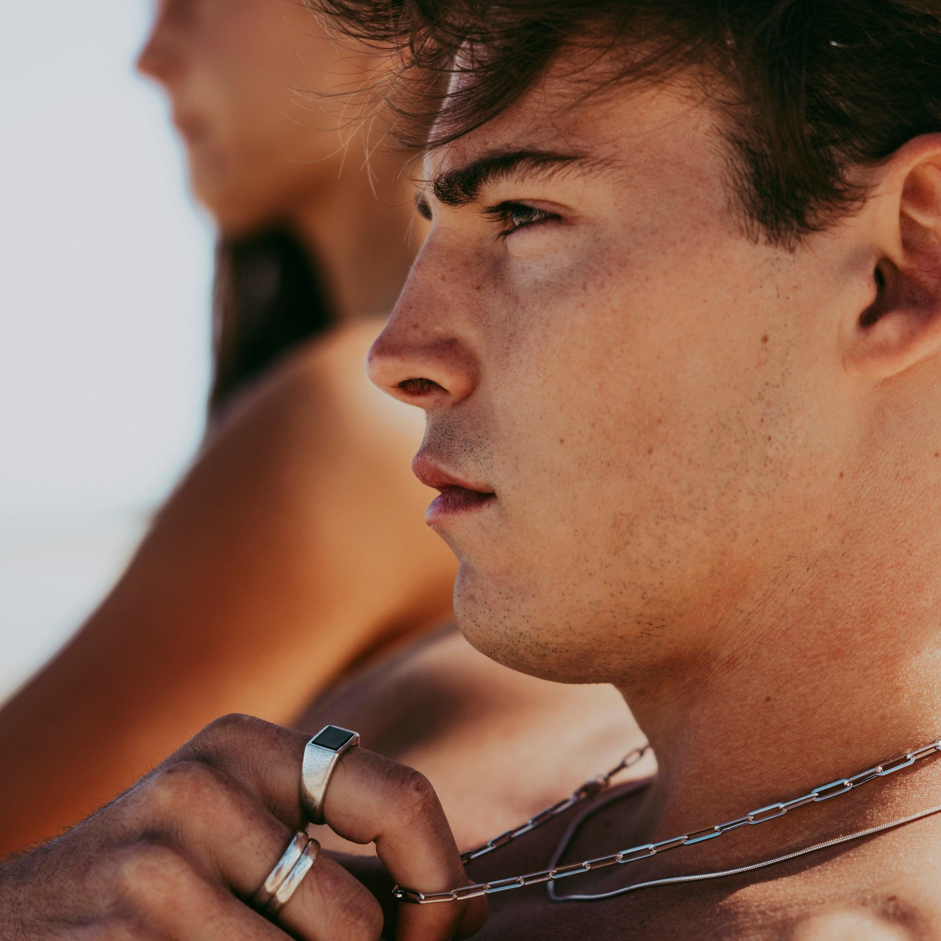 A man with a thoughtful expression adjusts his layered necklaces, including the Billie Jo Snake Necklace. The image captures a moment of quiet confidence and style.