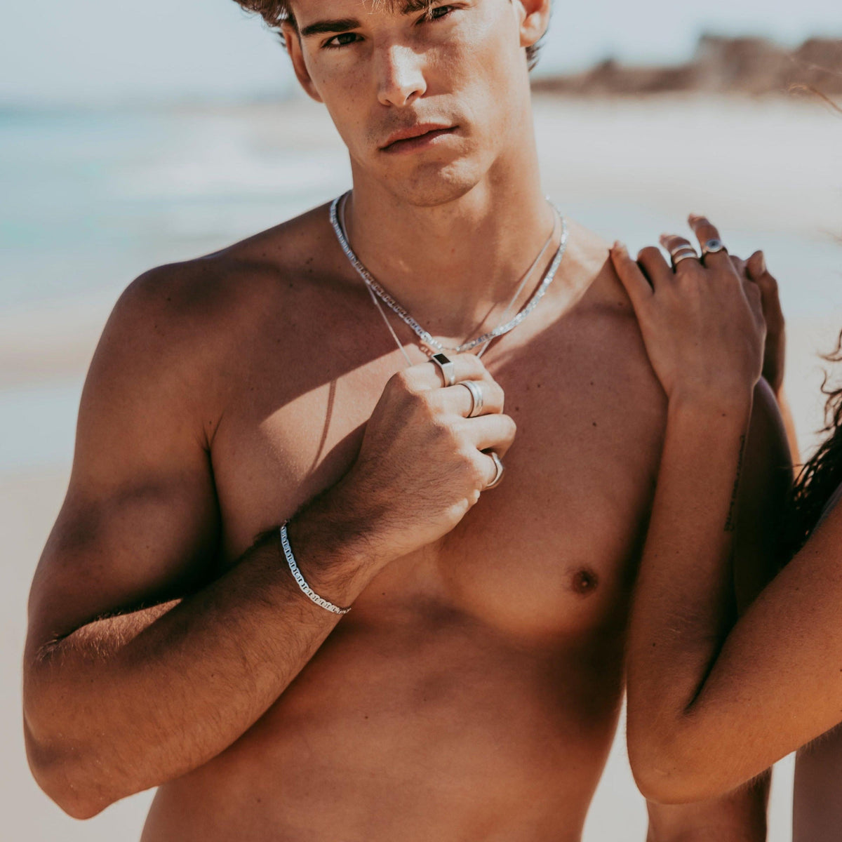 A man on the beach wearing the sterling silver Mariner Bracelet from Billie Jo, with a woman's hand on his shoulder. A summer lifestyle shot.