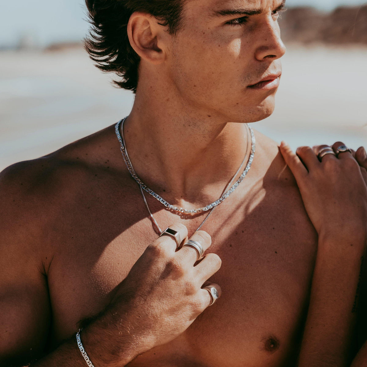 A male model wears the silver Mariner Necklace from Billie Jo, layered with other chains and rings for a stylish, beachside editorial look.