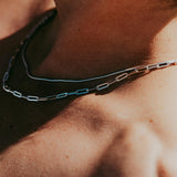 A man wears the sterling silver Snake Necklace from Billie Jo, layered with another chain. The close-up shot highlights the necklace's texture against his skin in the warm sunlight.