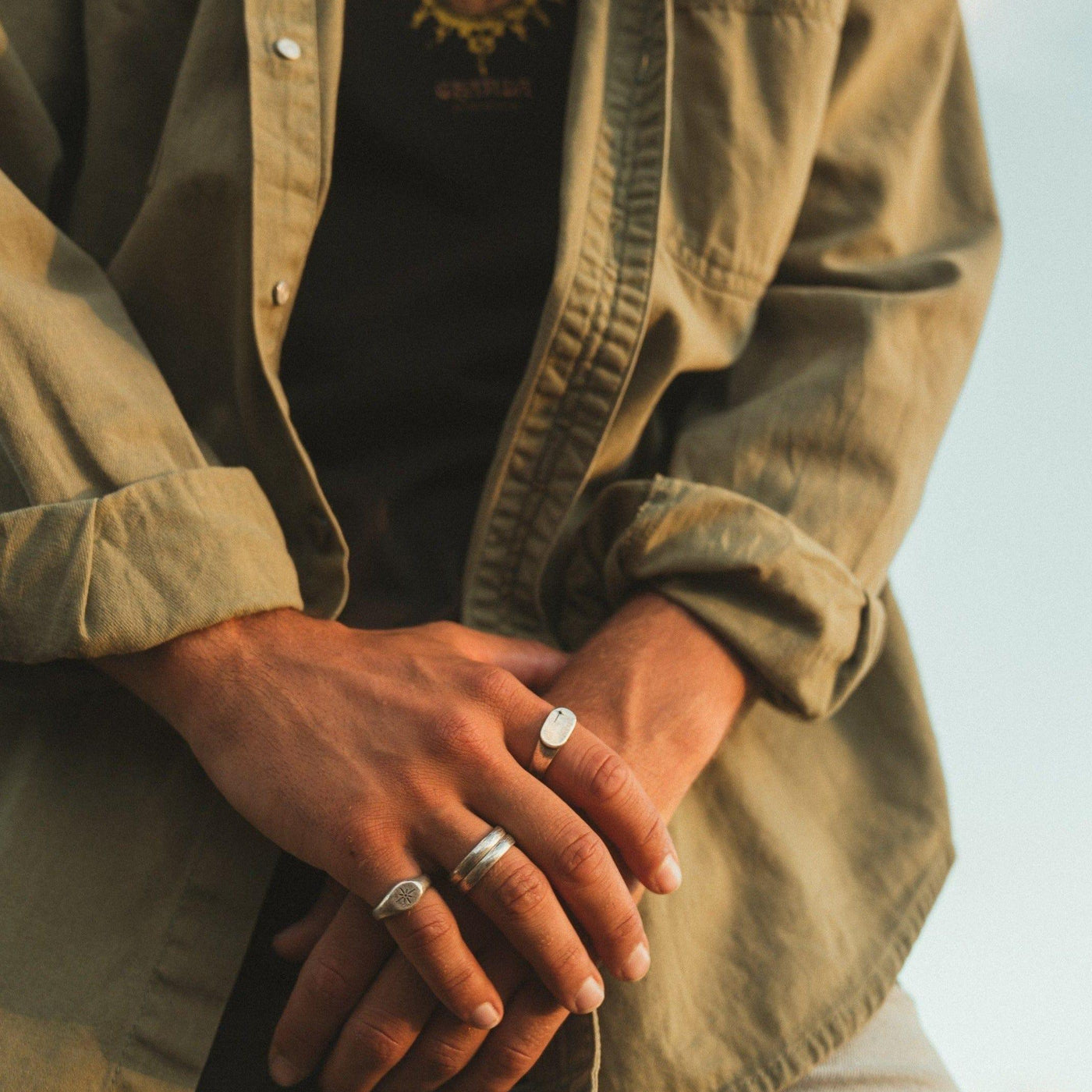 A man wearing a stack of silver rings, including the Billie Jo Luminous Ring, with a casual, layered shirt. An editorial shot for the Australian men's jewellery brand.