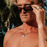 A man with sunglasses wears the Billie Jo Untold Pendant, a sterling silver necklace with a round, textured pendant, in a sunny, outdoor setting.